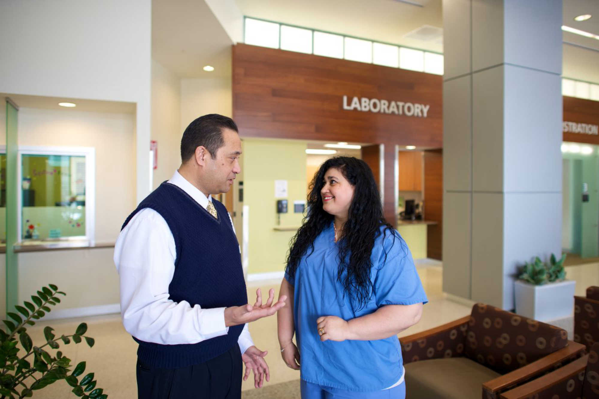 A female medical professional in scrubs discusses work with her male coworker who is dressed in business professional attire at a health clinic.